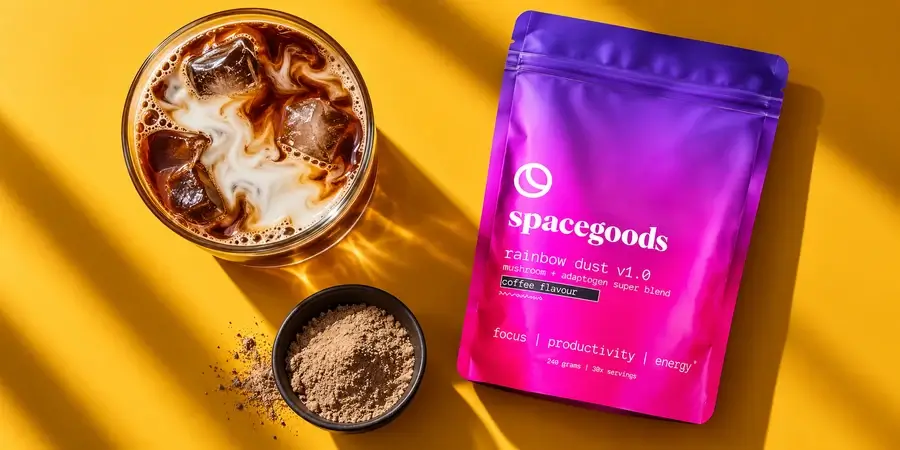Glass of iced coffee with milk next to a bag of Spacegoods Rainbow Dust and a bowl of powder on a mustard background