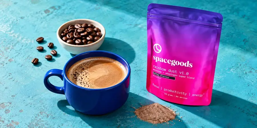 Bag of Spacegoods Rainbow Dust beside a blue mug of coffee and a bowl of coffee beans on a teal background