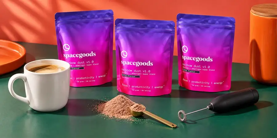 Three bags of Spacegoods Rainbow Dust behind a mug of coffee, a scoop of powder and a small milk frother against a coral wall